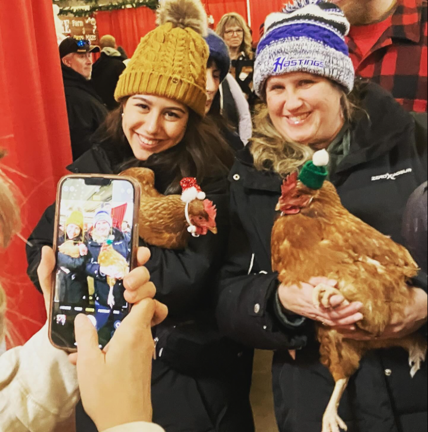 Two light-skinned women in winter gear hold chickens, each of which is wearing a small Santa hat, as they smile and pose for a photo.