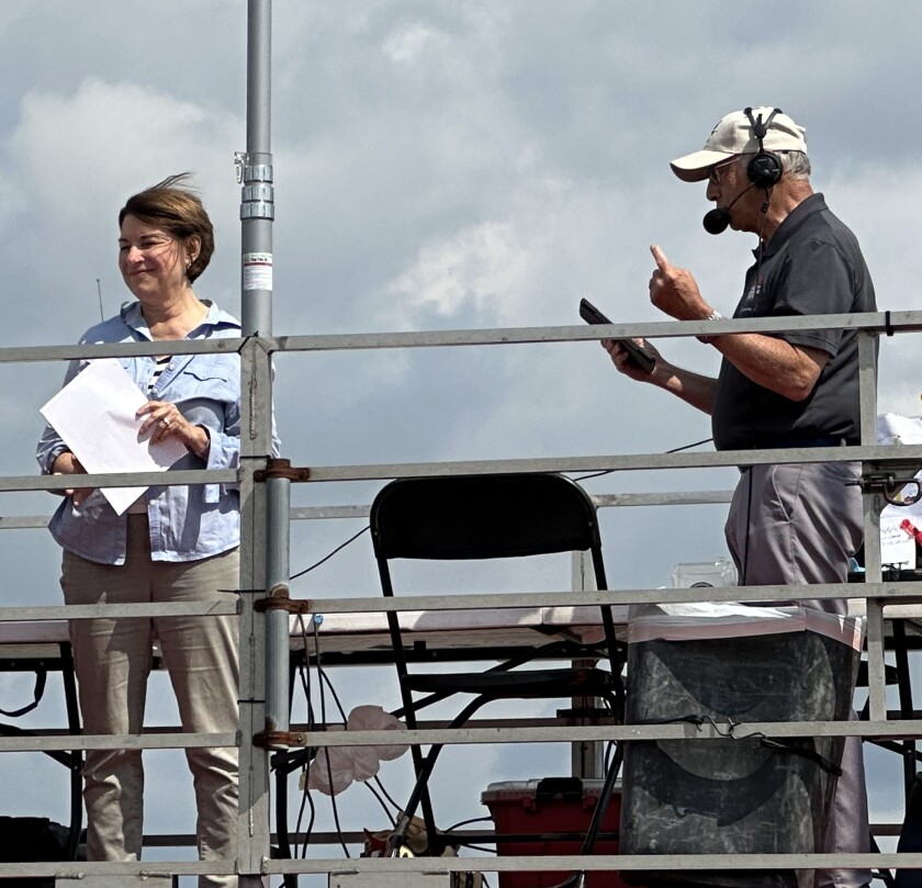 On an announcer stand bounded in a high metal railing, a light-skinned woman holds a sheet of paper and smiles as a light-skinned man speaks into a microphone on his headset.