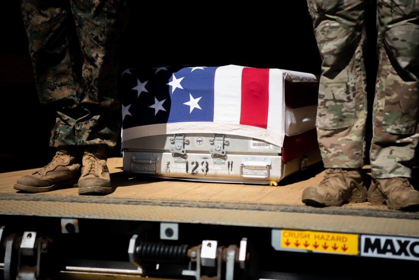 Two service members stand next to a flag-draped casket or transfer case