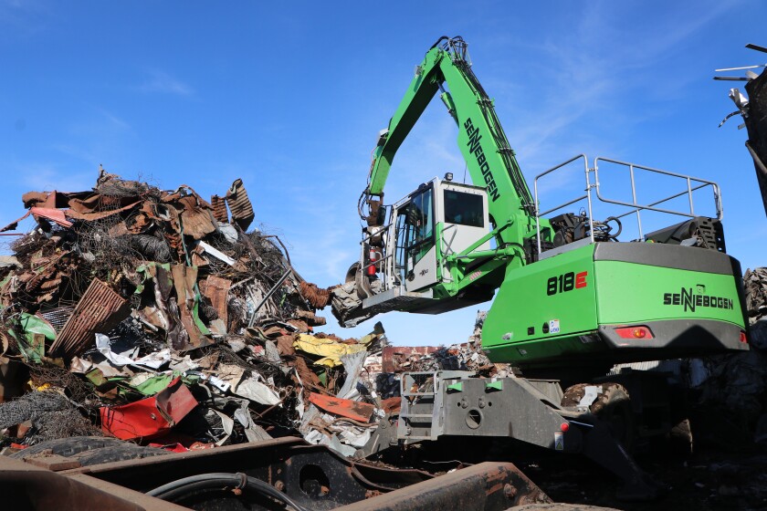 A green German-made materials mover picks up scrap iron to be placed in a truck. The cab has bulletproof glass and can be lowered or raised, hydraulically from about 10 feet to 18 feet, with 360-degree movement.