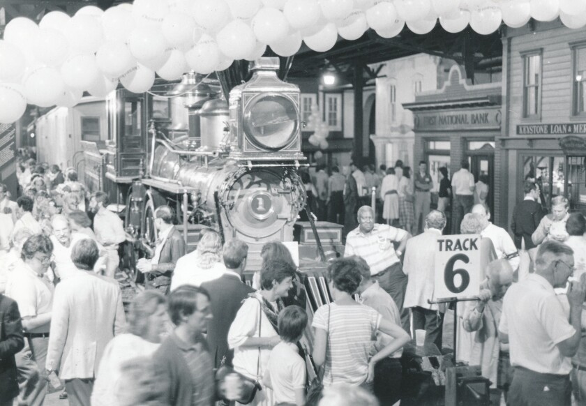 In a black-and-white image, a dense crowd of people surround a steam locomotive with a foreground sign reading "TRACK 6."