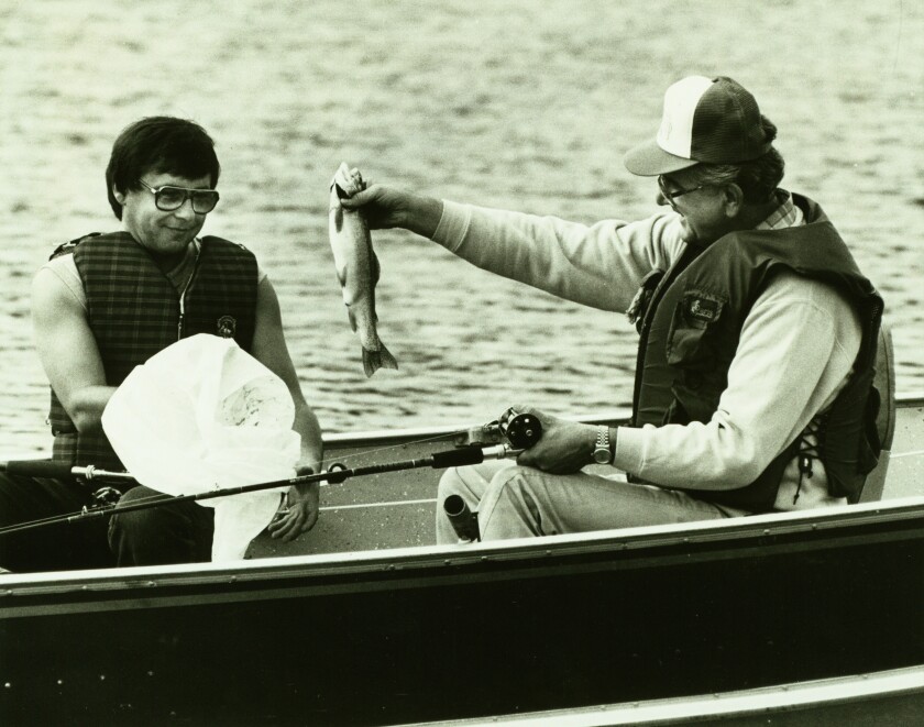 Two men sit in a boat, fishing pole at the ready. The man to the right holds up a fish, while the man to the left smiles and seems to hold a bag open to receive it.