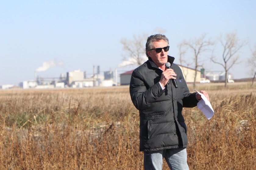 A man in a black coat, jeans and sunglasses speaks with a microphone, flanked by an ethanol plant in the distance.