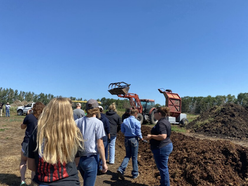 A woman dressed in a black polo shirt and blue jeans stands in front of a manure compost heat and talks to a group of youth.