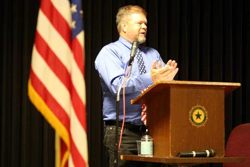 A man clapping while standing at a podium near an American Flag and giving a speech at a Veterans Day event.