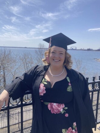A woman in a graduation cap and gown smiles with Lake Superior and the Aerial Lift Bridge behind her.