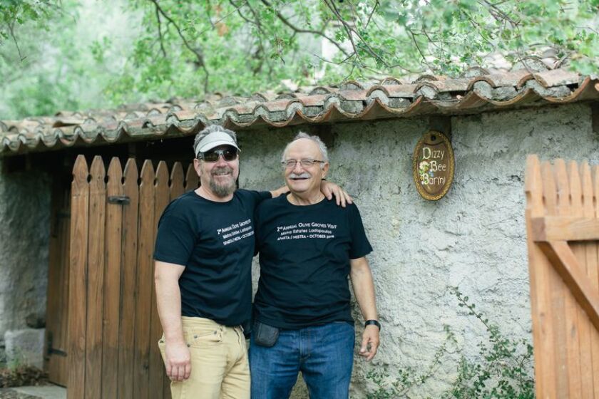 Two men with arms around each other's backs stand in front of a stone building.