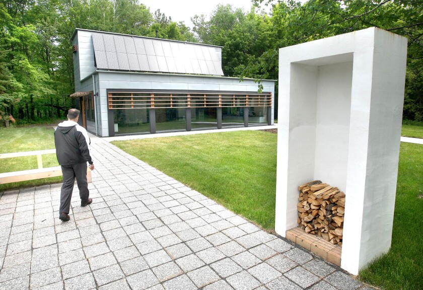 Light-skinned man walks across a granite patio towards a low modern building bedecked with solar panels, with firewood stacked in high rectangular structure in foreground at right.