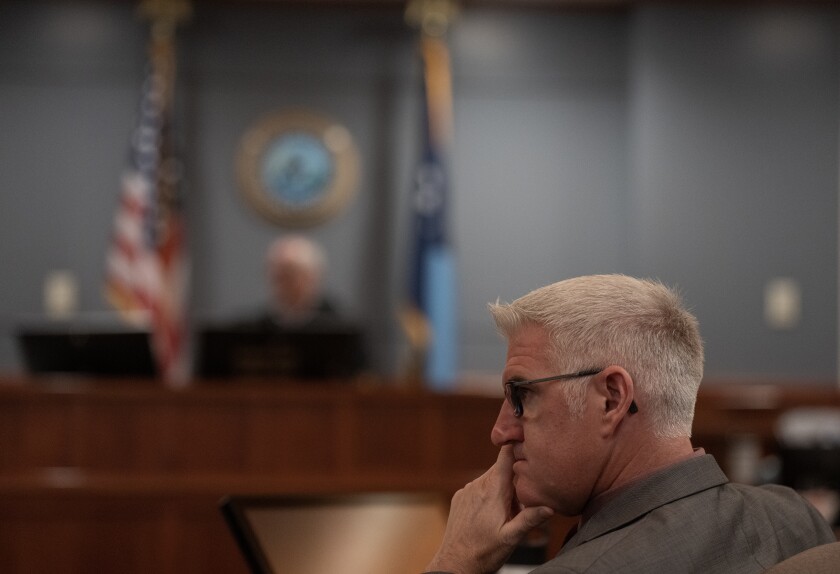 A white-haired man pensively holds his chin and looks to the side. Blurred in the background, a judge sits at the head of a courtroom.