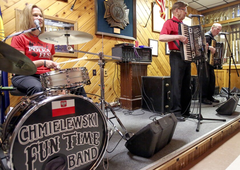 Three white polka musicians play on a low stage in front of a wood-panel wall. Head of bass drum, in foreground, reads, "Chmielewski Funtime Band."