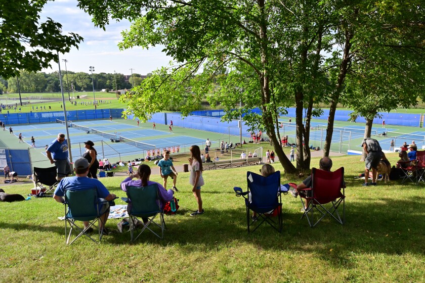 Fans sit in lawn chairs in the shade from trees on the hillside overlooking the tennis courts.