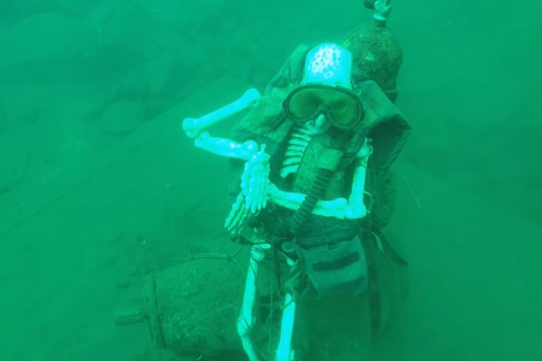 A skeleton dressed in scuba gear sits on a green lake bottom under water.