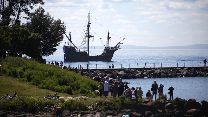 Tall ship moving in the water while people watch from shore.