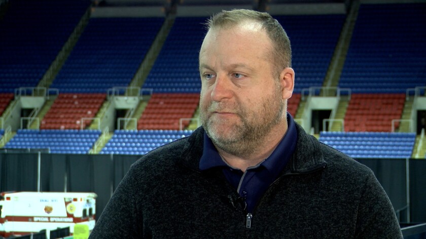 David Ripplinger, in a black fleece pullover and a blue polo shirt stands in front of bleachers.