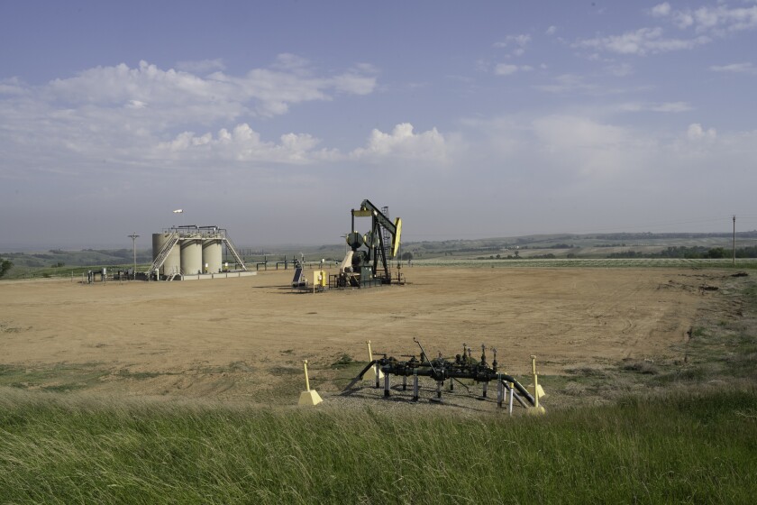 An oil derrick stands in a clearing on the plains, with several large tanks in the background and some pipes in the foreground