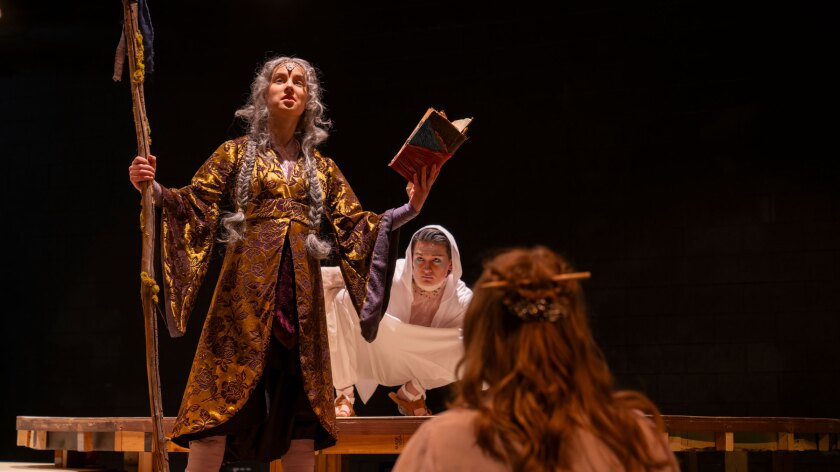 In a black-box onstage setting, a person clad as a golden-gowned sorcerer stands at center, holding a staff and a book. A white-clad person crouches behind.