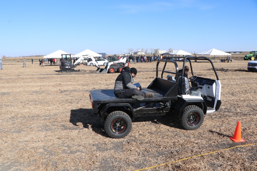 Workers prepare a white, small, autonomous truck for a demonstration, as a groundbreaking crowd listens to a program in the background.