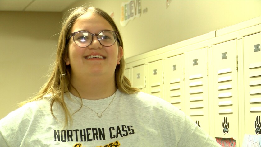 A girl in glasses with her hair down smiles toward the camera. She is wearing a t-shirt.
