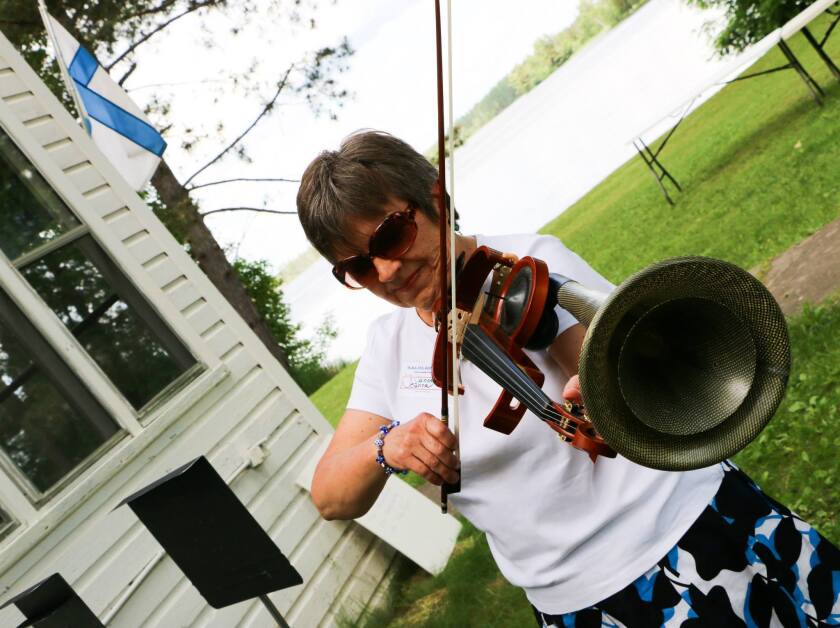 A woman plays a violin with amplifying horn attached. Visible in the background are a lake and a white wooden structure flying a Finnish flag.