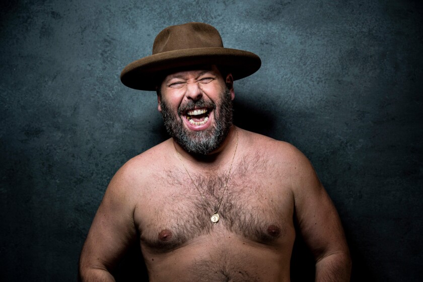 Hairy white man with bare torso grimaces playfully while flexing arms, wearing brown hat and metal necklace.
