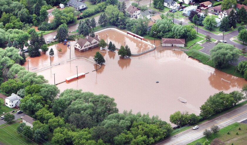 A rectangular park seen from the air, surrounded by trees, is under brown water, with the tops of soccer nets, the edges of hockey rinks, and a fieldhouse visible