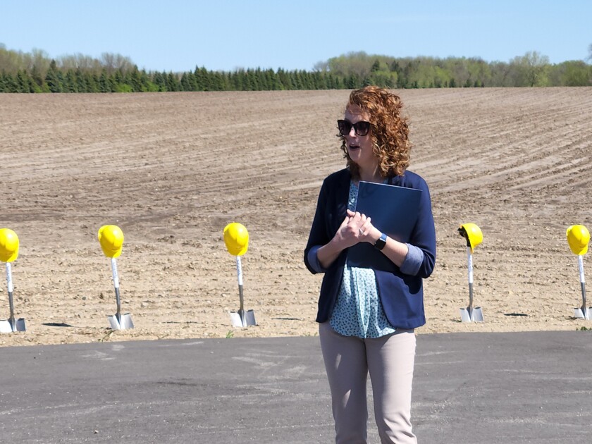 A woman stands in front of a dirt covered field and shovels topped with yellow hard hats speaking to attendees of a groundbreaking ceremony.