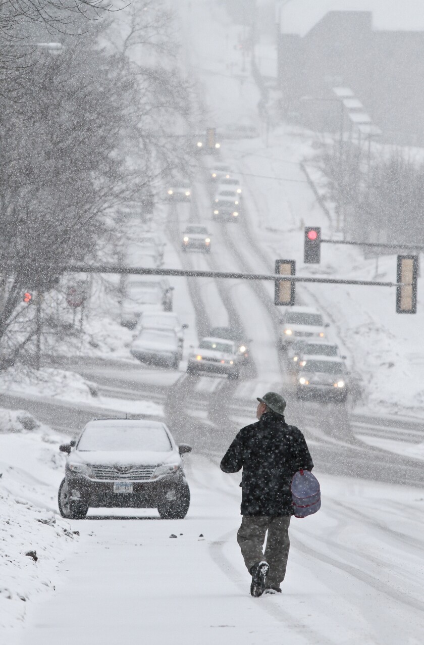 Cars travel down a steep hill in snowy conditions, stopping at a traffic signal. A pedestrian walks in the foreground