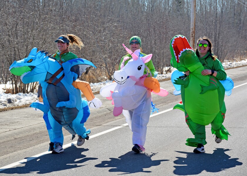 Three runners on street, wearing fantastical animal costumes: a dragon, a unicorn, and a dinosaur.