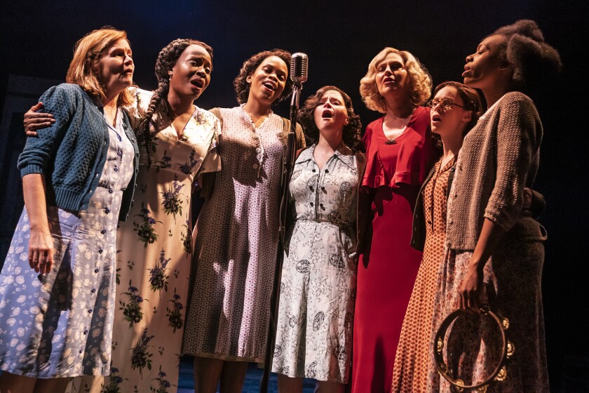 Seven women of varying skin colors, dressed in modest 1930s garb, stand around a microphone, singing passionately with their eyes closed.