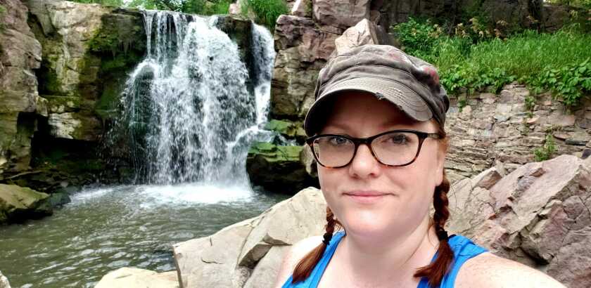 Woman in pigtails and a baseball cap smiles for the camera with a waterfall behind her.