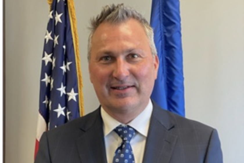 light-skinned man wearing gray suit and blue tie, standing in front of two flags