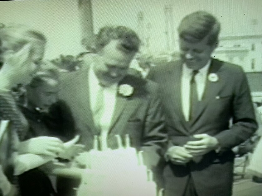 John F. Kennedy stands next to a man in a suit looking at a birthday cake.