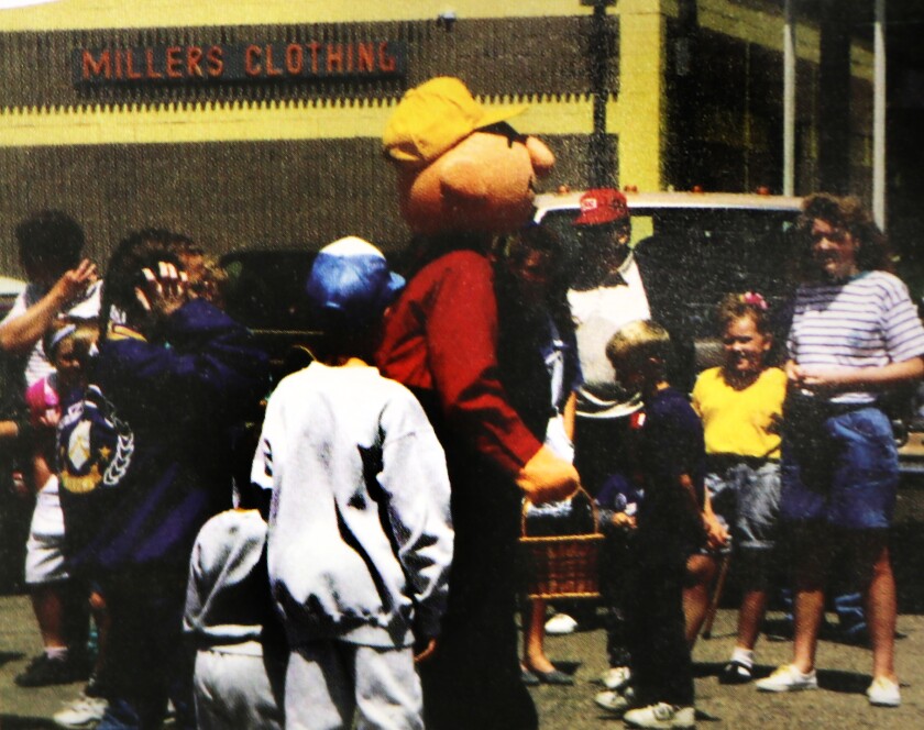 A cartoon character, looking like a round-faced farmer in a yellow cap, strides through a group of children in a street parade.