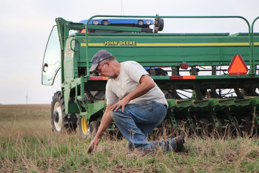 A farmer takes a knee to evaluate soybean seed placement during planting, flanked by a no-till John Deere planter.