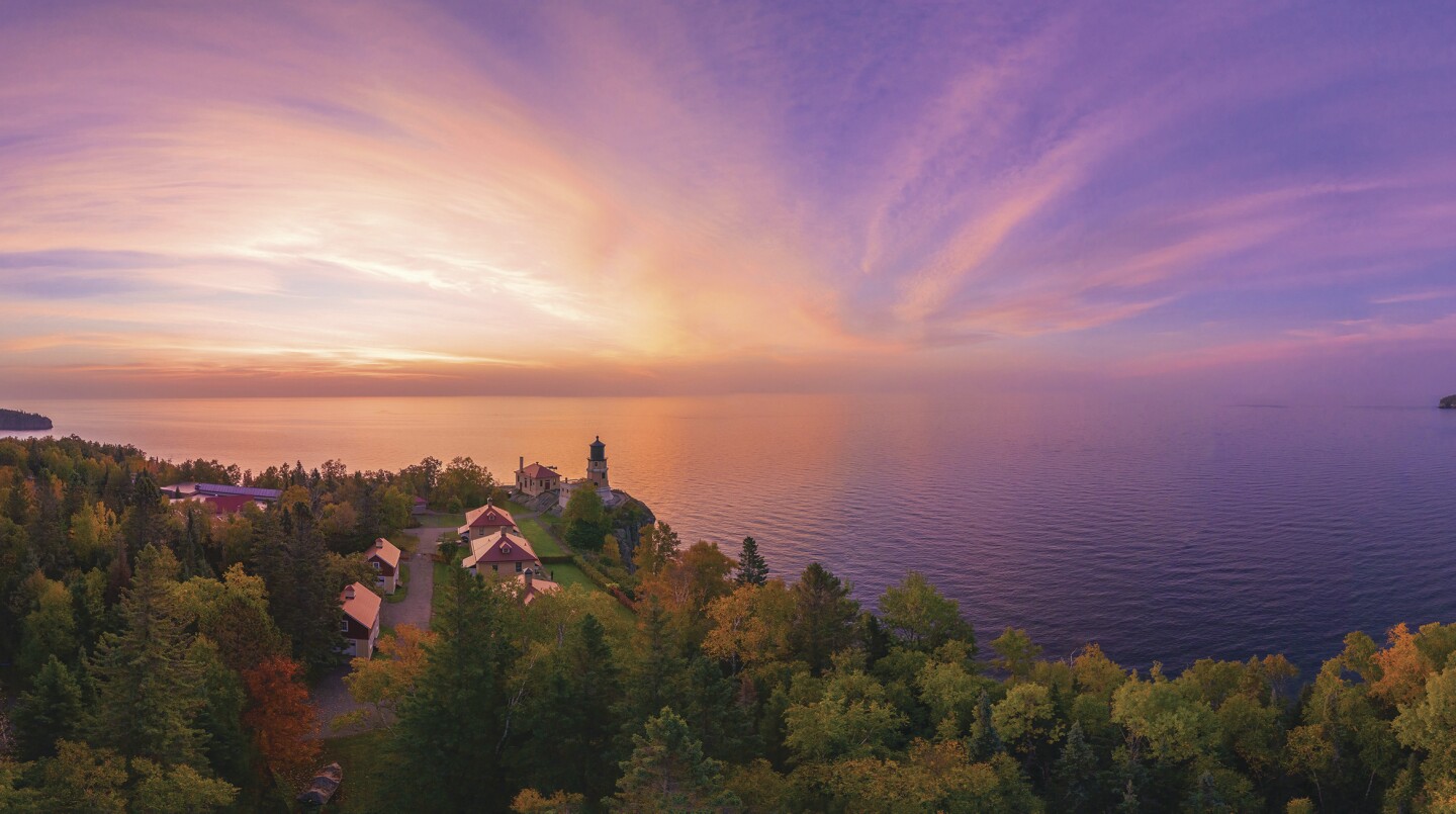 Sun rises over Lake Superior, with color fading from white to a purplish blue, in an aerial photo. Tree-lined shoreline with lighthouse seen in foreground.