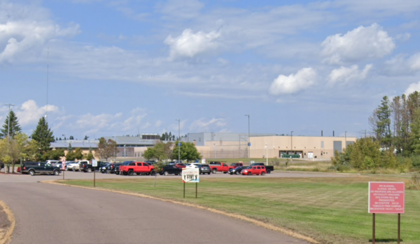 A photo shows a series of low buildings surround by a fence and trees