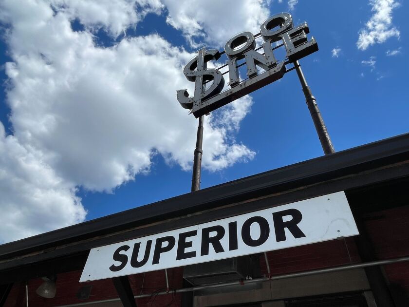 A daylight view from a low angle up at a depot building with a sign reading SUPERIOR in bold letters. A neon sign reading SOO LINE rises above the building, with fluffy clouds visible in the sky.