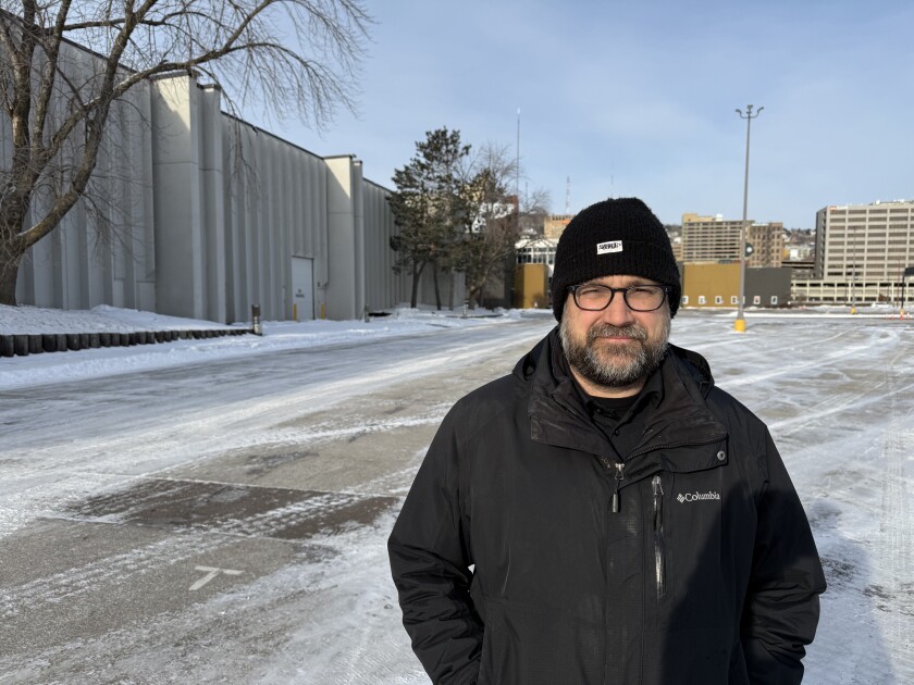 White man wearing black winter garb stands in empty parking lot in front of blocky white building, with hillside cityscape in background.