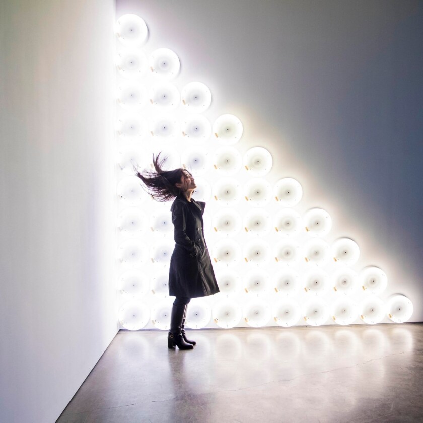 A woman dressed in a black coat and boots sands smiling in front of a sculpture with bright white lights.