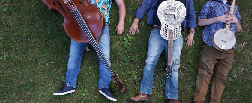 Overhead photo of three men lying on their backs in the grass, stringed instruments laid over their bodies, their heads cropped out of the photo.