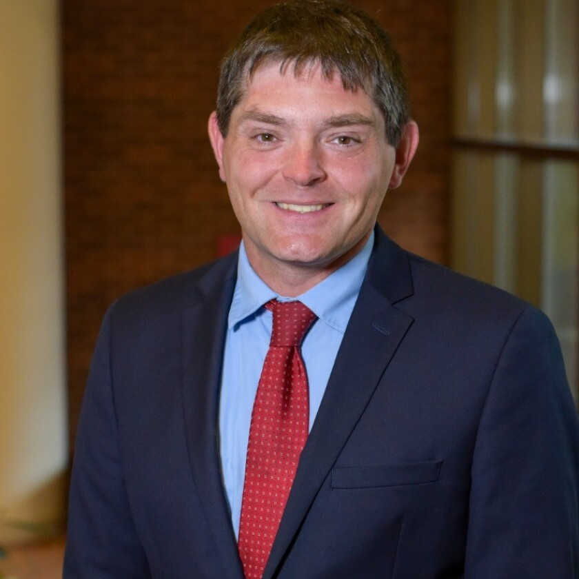 A white man with short, brown hair smiles for a portrait. He wears a dark suit with a red tie and light blue shirt.