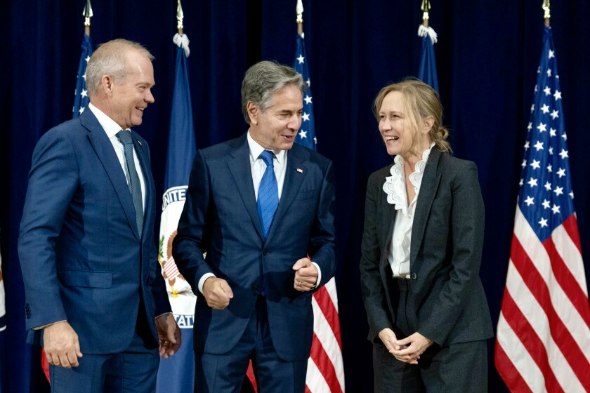 From left, all white and wearing suits, a man with light gray hair, a man with salt-and-pepper hair and a blond woman chat in front of several United States flags.