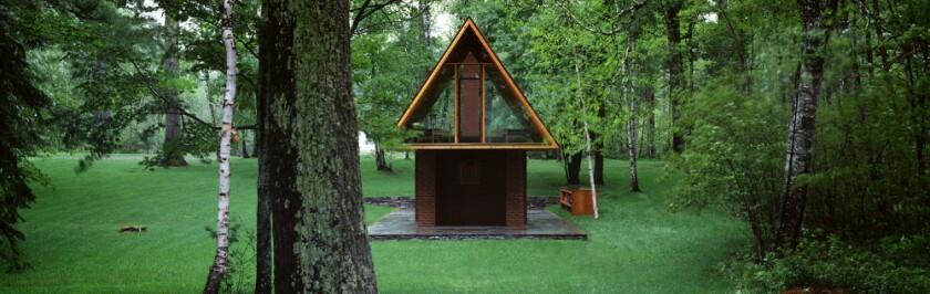 Small building with brick volume containing concave curved wall supporting wooden, triangular upper level, located in a verdant wooded area.