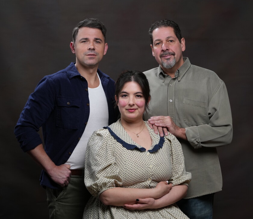 Three people pose together for portrait in front of dark backdrop. All are dressed simply, with two men at rear wearing button-down shirts and woman in front wearing dress redolent of the early 20th century.