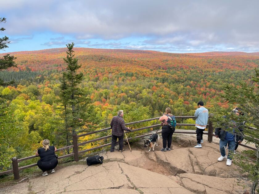 Several people, including one holding a dog on a leash, gather against a wooden fence and enjoy a wide view of fall colors from a tall, wide rock.
