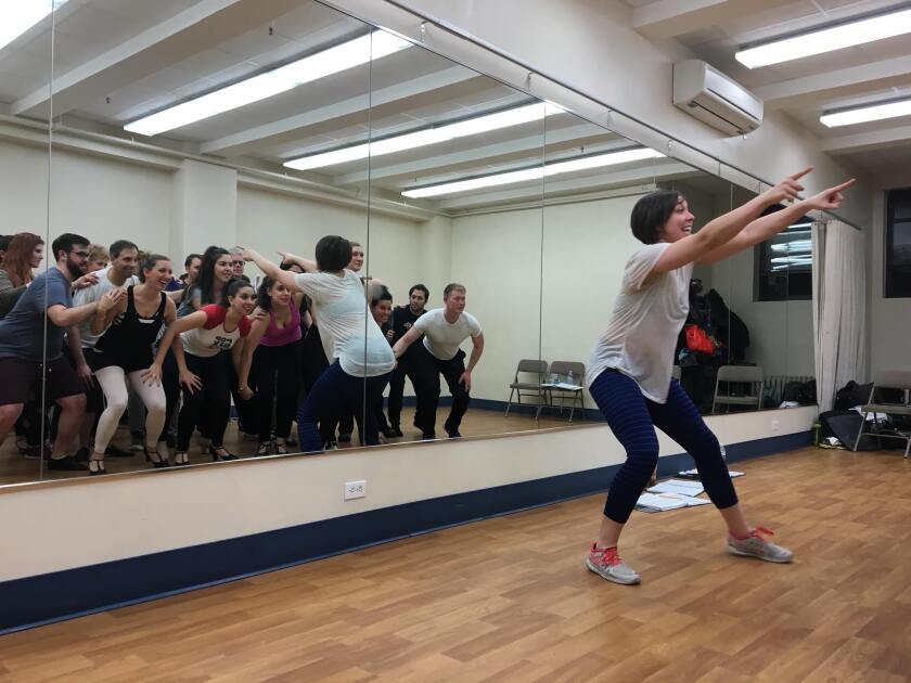 Woman in workout wear is shown in a rehearsal room. Through a mirror at left, it can be seen that she's pointing at a large cluster of smiling dancers.