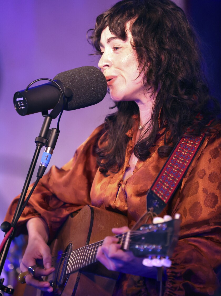A light-skinned woman with dark, curly hair plays acoustic guitar and sings into a microphone.