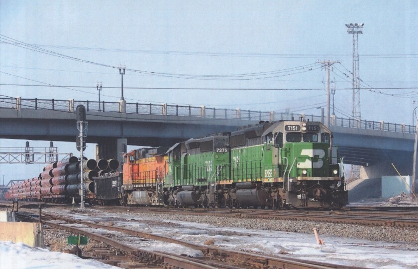 Diesel locomotive at the head of a freight train on tracks passing beneath a bridge. Locomotive is painted green, with "BN" logo.