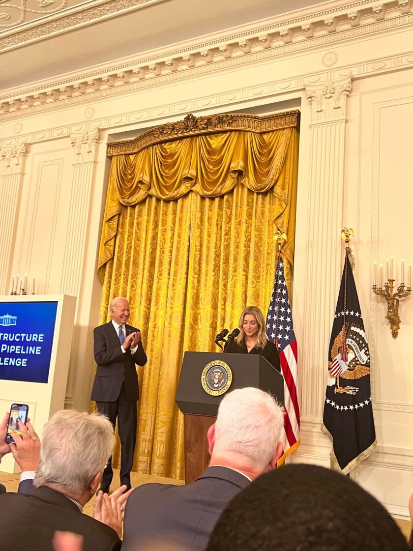 President Joe Biden claps as a woman speaks at the podium before a group.