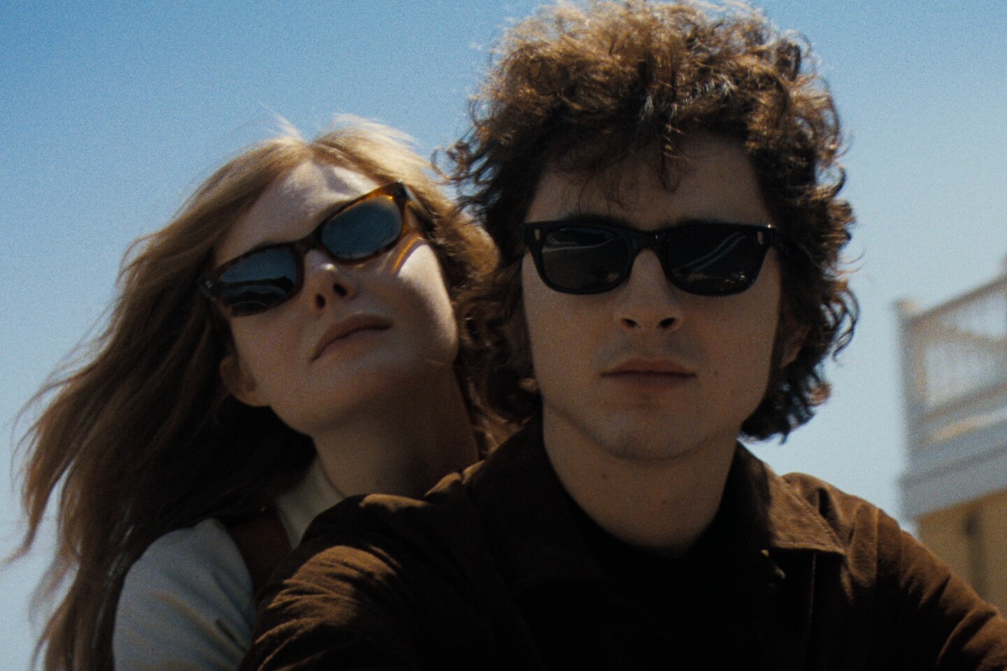 White woman and man in their 20s seen in close-up, wind blowing in their hair. Both are wearing sunglasses, and a low yellow building is seen in the background.